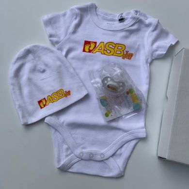 Babyset mit weißem Body, Mütze und Schnuller, alle mit ASBaby-Logo; daneben liegt eine weiße Verpackungsschachtel.