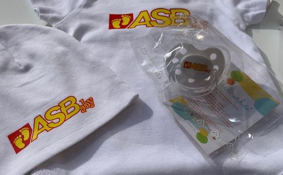 Baby-Erstausstattung mit ASB-Logo: weiße Mütze, Body und Schnuller im Set, alle mit rotem ASBaby-Schriftzug und gelben Füßchen.