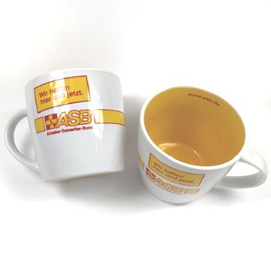 Zwei weiße Tassen mit gelber Innenseite, ASB-Logo und Slogan „Wir helfen hier und jetzt“, eine liegt, eine steht.