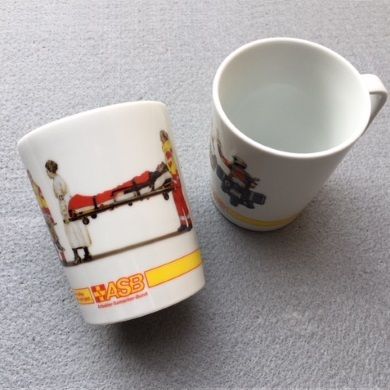 Zwei weiße Tassen mit ASB-Logo, eine zeigt Rettungskräfte mit Trage, die andere steht aufrecht, beide auf grauem Untergrund.