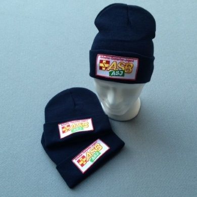 Zwei dunkelblaue Mützen mit ASJ-ASB-Logo, eine auf einem weißen Schaufensterkopf, die andere…
