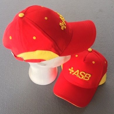 Zwei rote ASB-Kappen mit gelben Elementen und gesticktem Logo, eine auf Styroporkopf, eine flach auf dem Boden liegend.