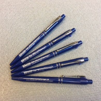 Fünf dunkelblaue Kugelschreiber mit silberner Aufschrift „Der Wünschewagen“ und Website, fächerförmig auf grauem Untergrund.