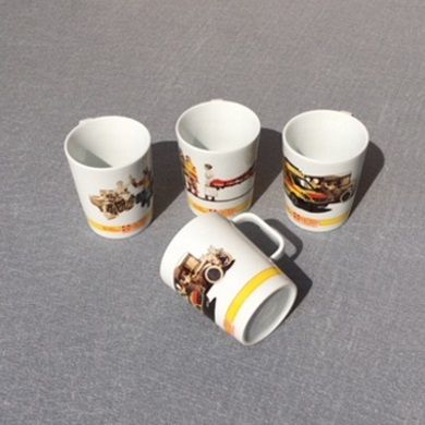 Vier weiße Tassen mit ASB-Design und Fahrzeugmotiven, drei stehen aufrecht, eine liegt seitlich auf grauer Oberfläche.