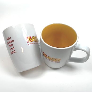 Zwei weiße Tassen mit gelber Innenseite, ASB-Logo und rotem Schriftzug „Wir helfen hier und jetzt“, eine stehend, eine liegend.