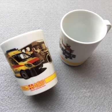 Zwei weiße Tassen mit ASB-Logo und Rettungswagenmotiv, eine steht aufrecht, die andere liegt seitlich auf grauem Untergrund.